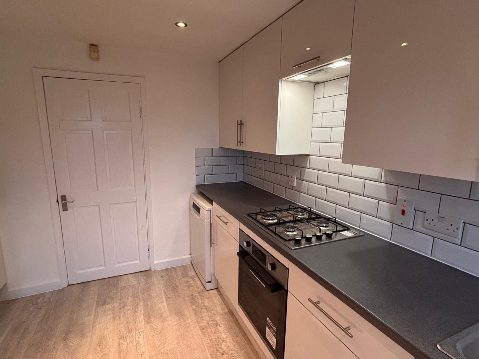 Refurbished flat kitchen completed by CJD &mdash; white shaker-style units, dark grey worktop, white metro-tile splashback, integrated oven, four-ring gas hob and washing machine, wood-effect vinyl flooring.