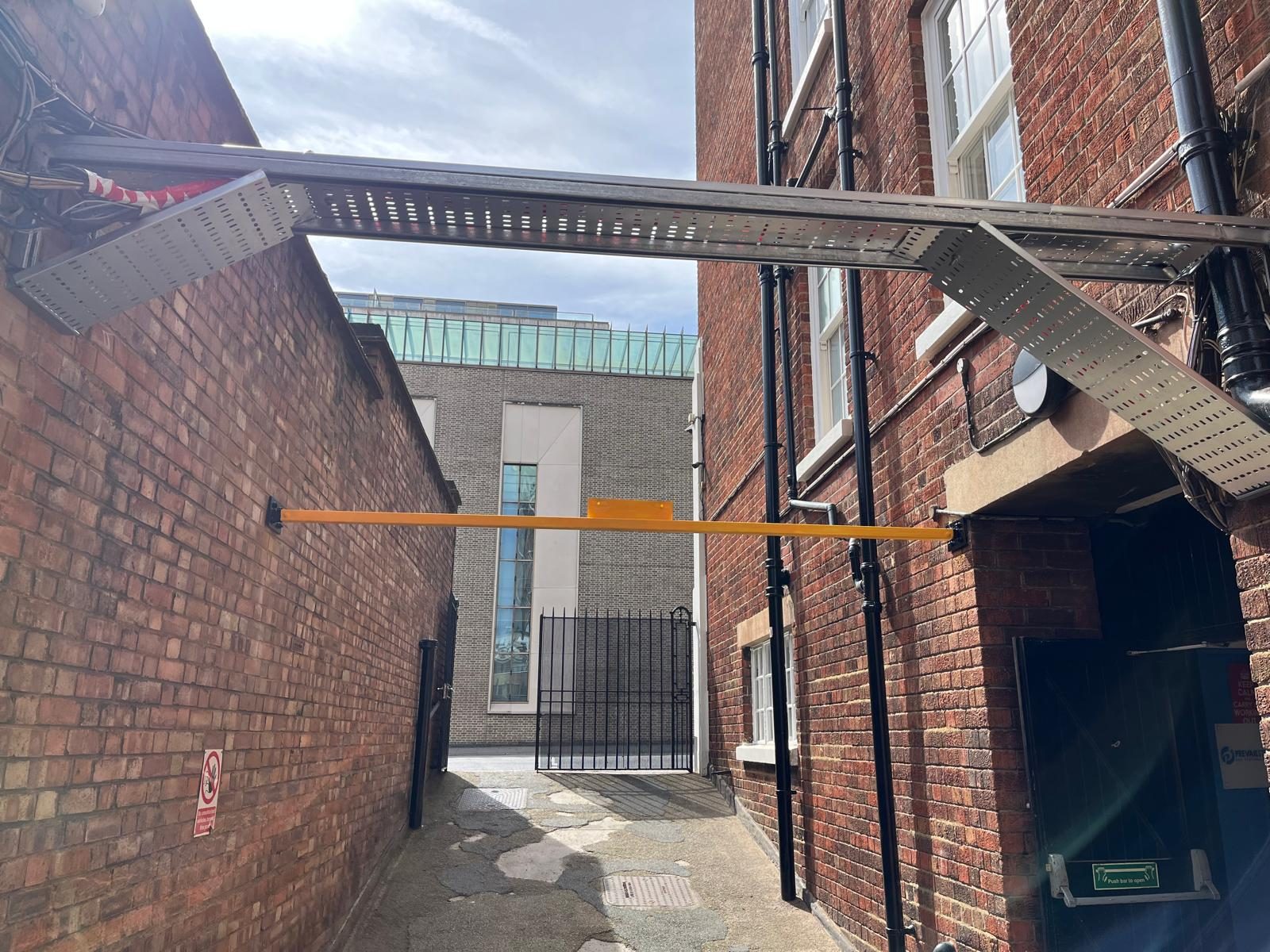 Galvanised steel cable tray and painted yellow structural beam spanning a narrow alleyway between two red-brick London blocks &mdash; external steelwork supplied and installed by CJD.